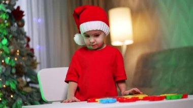Yaratıcı festival oyun zamanı! Kırmızı tişörtlü ve Noel Baba şapkalı bir çocuk, Noel süsleri arasında renkli oyuncaklarla oynuyor..