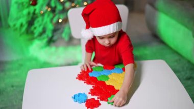 Çocuk yapboz yapıyor. Kırmızı tişörtlü ve Noel Baba şapkalı bir çocuk, şenlikli dekorasyonların ortasında beyaz bir masada bir yapboz oluşturuyor..