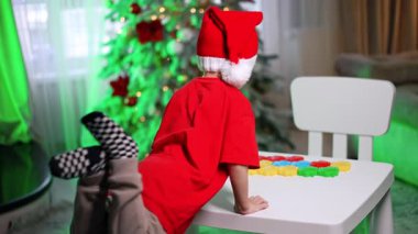 Çocuk ağaçla şekillerle oynar. Şenlikli şapkalı bir çocuk, Noel süsleri ve bir ağaçla çevrili bir masada renkli şekiller düzenler..