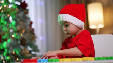 Ağacın yanında blokları olan çocuk. Kırmızı şapkalı küçük bir çocuk süslü bir Noel ağacının yanında renkli yapı taşlarıyla oynuyor..