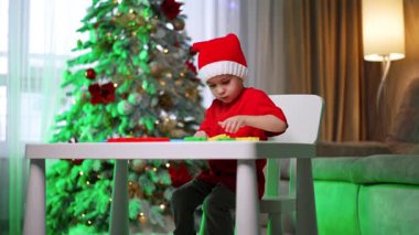 Çocuklar tatillerde oyun oynamaya bayılır! Şenlikli şapkalı bir çocuk dekore edilmiş bir Noel ağacının yanında yaratıcı bir şekilde bir masa inşa eder..