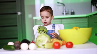 Çocuk vejetaryen yemeği yapıyor. Parlak bir mutfakta çeşitli taze sebzelerle çevrili bir masada oturmuş brokoliyi inceliyor..