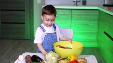 Çocuk sebzeleri yemek için hazırlıyor. Tulumlu bir çocuk yeşil ışıklandırmalı modern bir mutfakta yemek pişirirken bir kasede taze sebze topluyor..