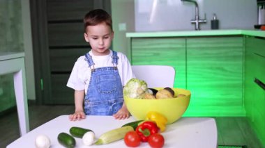 Çocuk içeride sebze ayırıyor. Modern bir mutfağın masasında renkli sebzeleri sınıflandıran tulumlu genç bir çocuk oyuncu merakını gösteriyor..