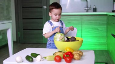 Çocuk mutfaktaki sebzelere bakıyor. Genç bir çocuk, çağdaş bir mutfaktaki masanın başında duruyor ve bir kasedeki çeşitli taze sebzeleri izliyor..