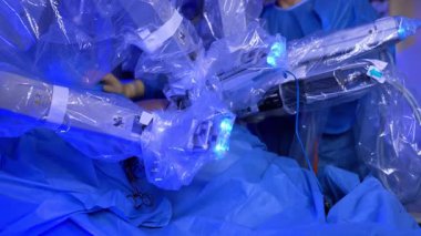 Da Vinci cerrahı, sterilite için plastikle kaplanmış robotik kollar. Mavi bornoz giyen doktor fütüristik makinenin önünde duruyor..