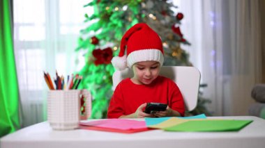 Kırmızı Noel Baba şapkalı sevimli çocuk masada oturuyor. Küçük çocuk elindeki akıllı telefona odaklanmış, tuşlara basıyor, arama yapıyor..