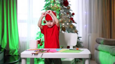 Komik çocuk Noel Baba şapkasını yüzüne çekiyor. Bebek renkli kalemler ve kağıt parçalarıyla masaya oturur..