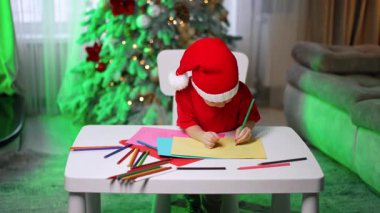 Kırmızı tişörtlü ve Noel Baba şapkalı küçük bir çocuk evdeki küçük masada oturuyor. Renkli kalemlerle odaklanmış solcu çocuk çizimi.