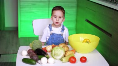 Beyaz tişörtlü ve kot pantolonlu beyaz bir çocuk masada oturuyor. Küçük çocuk sebzeleri mutfakta öğreniyor..