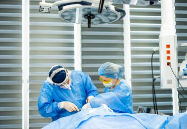 Modern ameliyathanede cerrah. Mavi önlüklü cerrahlar, hasta güvenliğini ve hassasiyetini sağlamak için parlak ışıklar altında hassas bir operasyon gerçekleştiriyorlar..