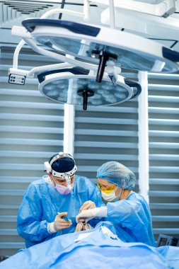 Cerrahlar karmaşık ameliyatlar yaparlar. Mavi önlüklü iki cerrah iyi donanımlı ve steril bir ameliyathanede bir hastayı ameliyat ediyorlar..