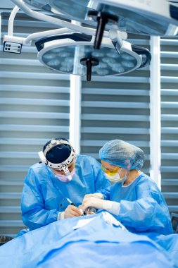 Cerrahi ekip ameliyat eder. Mavi önlüklü iki tıp uzmanı steril bir ameliyathanede ameliyat sırasında bir hastaya odaklanıyor..