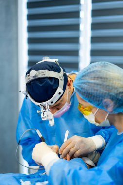 Hastane cerrahları. Mavi önlüklü ve maskeli iki cerrah modern bir hastane ortamında cerrahi müdahaleye odaklanmış durumdalar..