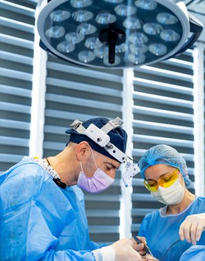 Cerrahlar hassas ameliyatlar yaparlar. Cerrahi önlüklü cerrahlar hastane ameliyathanesinde parlak ışıklar altında ameliyat yaparlar..
