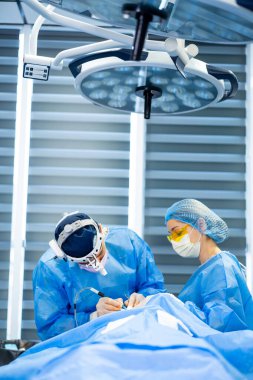 Tıbbi ekip tarafından ameliyat. Mavi önlüklü cerrahlar modern bir ameliyathanede parlak cerrahi ışıklar altında bir hastayı dikkatlice ameliyat ederler..