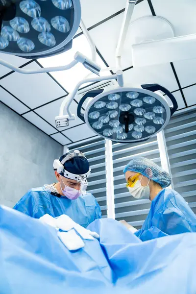 Cerrahlar ameliyathanede. İki maskeli cerrah steril bir ameliyathanede parlak ışıklar altında ameliyata konsantre oluyor..