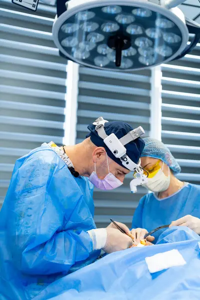 Cerrahlar ameliyathanede ameliyat eder. Koruyucu giysi giyen iki cerrah modern bir ameliyathanede parlak ışıklar altında cerrahi bir prosedüre odaklanıyor..
