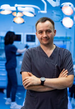 Doktor, asistanıyla ameliyathanede. Doktor, bir asistan arka planda cerrahi aletleri hazırlarken ameliyatta güvenle gülümser..