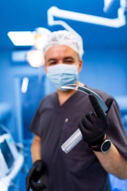 Cerrah ameliyathanede aletleri hazırlıyor. Önlüklü ve maskeli bir cerrah, bir laringoskop tutuyor. Parlak bir ameliyathanede ameliyata hazırlanıyor..