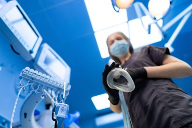 Doktor prosedüre hazır. Ameliyat maskesini önlüklü bir sağlık görevlisi tutuyor. Parlak bir ameliyathanede ameliyata hazır..