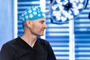 Cerrah ameliyata hazır. Önlüklü ve mavi şapkalı bir tıp uzmanı parlak bir ameliyathane düzenine odaklanmış durumda..