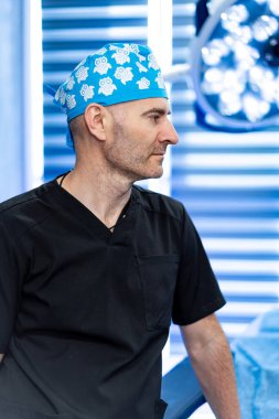 Cerrah ameliyathanede. Mavi şapkalı ve siyah önlüklü bir erkek cerrah parlak bir ameliyathanede ameliyata hazırlanıyor..