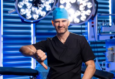 Cerrah ameliyathanede onaylamadığını gösteriyor. Ameliyat önlüğü ve kep takmış bir cerrah, parlak bir ameliyathanede tıbbi ışıkları olan bir jest gösteriyor..