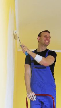 Çekici yakışıklı tamirci duvarı yeniliyormuş. İşyerindeki ressam, turuncu bir duvarı silindirle boyuyor. Dikey video