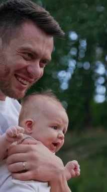 Beyaz tişört giyen sakallı adam kucağında bebek tutuyor. Sevgi dolu bir baba oğluna el sallıyor ve çocuğun başını öpüyor. Bulanık doğa arkaplanı. Dikey video