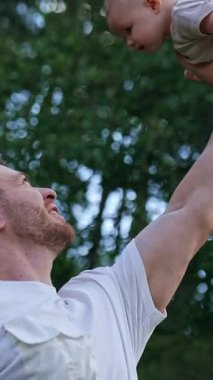 Kaslı beyaz bir adam tek eliyle yeni doğmuş bebeğini başının üstünde tutuyor. Gülümseyen mutlu baba oğlunu eğlendiriyor. Doğa arkaplanı. Dikey video