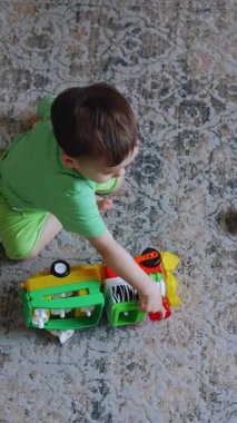 Bir yaşında bir çocuk odada halının üzerinde oturuyor. Siyah saçlı çocuk oyuncak trenle oynuyor. Üst Manzara. Dikey video
