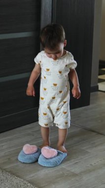 Sevimli bebek pofuduk terliklerin yanında duruyor. Çocuk evde giydiği ayakkabıları giyip halıya doğru koşuyor. Dikey video