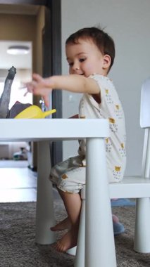 Tatlı beyaz bir erkek bebek masada oturuyor. Sevimli çocuk oyuncak hayvanlarla oynuyor. Düşük açı. Dikey video