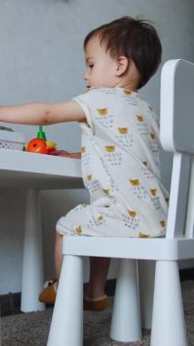 Beyaz elbiseli küçük çocuk masada oturuyor. Küçük çocuk parlak oyuncaklar oynar. Düşük açı. Dikey video