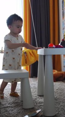 Küçük çocuk sandalyeleri masadan kaldırıyor. Bebek masadaki oyuncaklara dokunuyor ve sonra yere iniyor. Dikey video