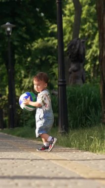 Aktif Kafkasyalı bebek parkta topla oynuyormuş. Çocuk oyuncağı fırlatıyor ve yolda mutlu bir şekilde koşuyor. Dikey video
