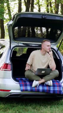 Düşünceli beyaz adam laptopuyla doğada çalışıyor. Mutlu adam arabanın bagajında oturur, etrafa bakar, fikirleri düşünür. Dikey video