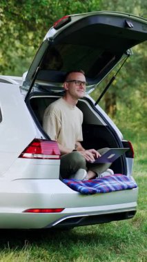 Mutlu adam ormanda arabasının bagajında oturuyor. Gülümseyen serbest çalışan biri dizüstü bilgisayarla doğanın tadını çıkarıyor. Dikey video