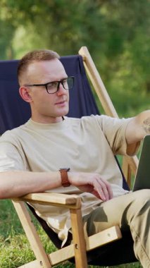 Kafkasyalı serbest yazar dizüstü bilgisayarla çalışmaya başlıyor. Adam çayırdaki sandalyede oturan dizüstü bilgisayarı açıyor. Araba arka planda. Dikey video