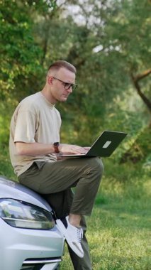 Beyaz bir erkek arabanın önünde dizüstü bilgisayar tutuyor. Bilgisayarda serbest çalışan adam tipleri ve gülümsemeler. Dikey video