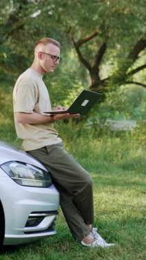 Orta yaşlı bir adam dışarıda arabaya yaslanıyor. Adam dizüstü bilgisayarı elinde tutuyor. Freelancer geleceğe bakarak gözlüklerini çıkartıyor. Dikey video