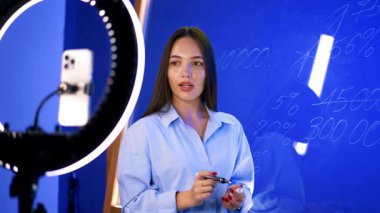 Esmer kadın hafif yüzükle kameraya konuşuyor. Kadın cam duvarda numaralar gösteriyor..