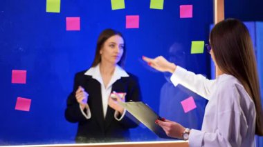 Cam duvarın önünde dikilmiş konuşan ve üzerine çıkartma yapıştıran iki kadın. İş arkadaşları yapışkan notlara işaret ederek iletişim kuruyor.