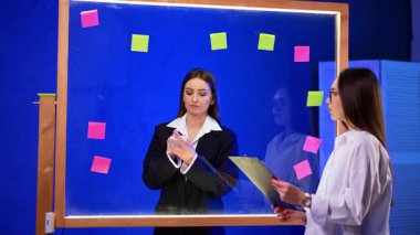 Uzun saçlı esmer kadınlar cam duvarın yanında dururlar. Kadın çıkartmalara yazı yazıp tahtaya yapıştırıyor. Planlama kavramı.