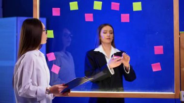 Elinde bir pano olan esmer, cam duvara bakıyor. Kurulun arkasındaki kadın üzerine etiket yapıştırıyor. Ofiste beyin fırtınası..