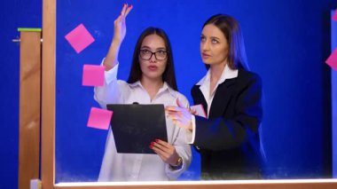 Bayan meslektaşlar cam duvarın yanında fikirler üzerinde çalışırlar. Kadınlar panoya ve tahtaya yapıştırılan etiketlere bakar..