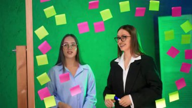 Hanımlar yapışkan notlara yazılmış fikirleri tartışırlar. Kadınlar cam duvardan notları alır ve geri atar..