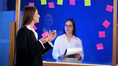 Cam duvarın yanında üzerinde numaralar ve çıkartmalar olan olumlu esmer konuşmalar var. Tahtanın arkasındaki kız elinde bir pano tutuyor..