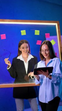 Gülümseyen beyaz kadınlar tablete bakarlar. Ofiste çalışan kadınlar cam duvarın önünde çıkartmalarla duruyorlar. Dikey video.